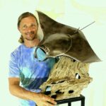 Carlos Hiller artist posing with his metalic sculpture of a manta ray.